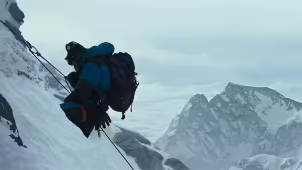 A climber in a blue down jacket ascends a steep icy slope on ropes