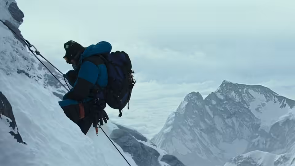 A climber in a blue down jacket ascends a steep icy slope on ropes