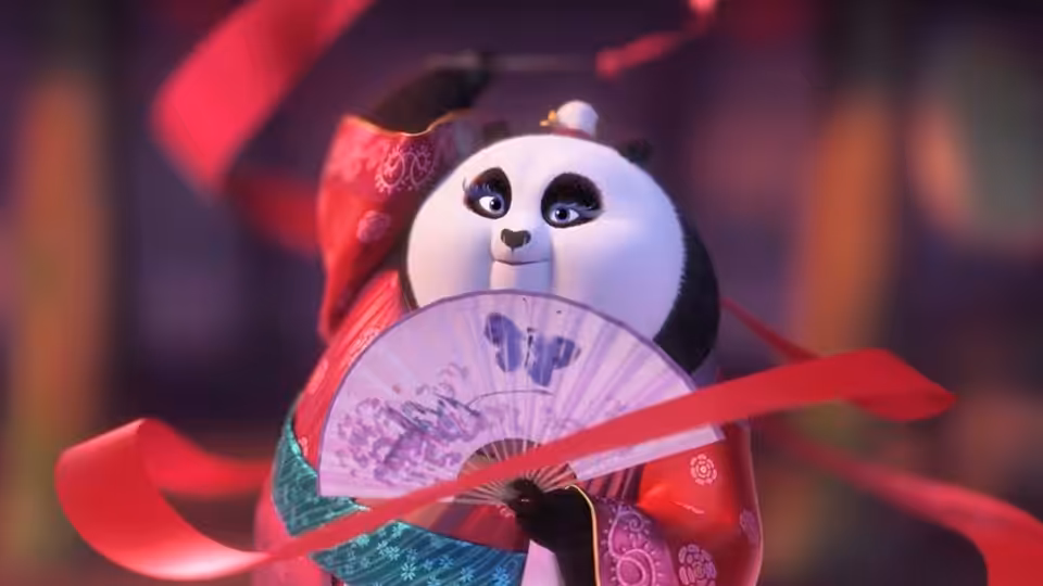 Po in a red robe holds an open fan across his face during a performance