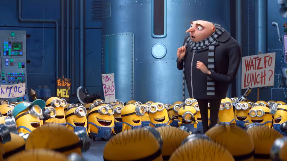 Gru addresses a crowd of Minions holding signs inside a blue industrial room