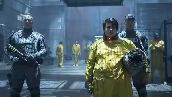 Firefist stands in a yellow jumpsuit between armed guards inside a prison