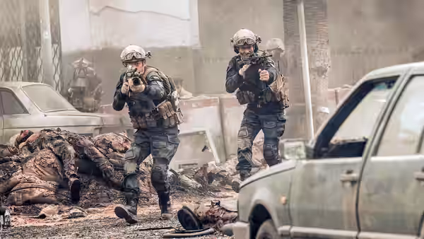 Two armed soldiers advance past bodies and abandoned cars on a rubble-strewn street