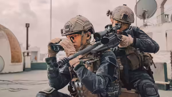 Two soldiers scan a rooftop through binoculars beside a scoped rifle