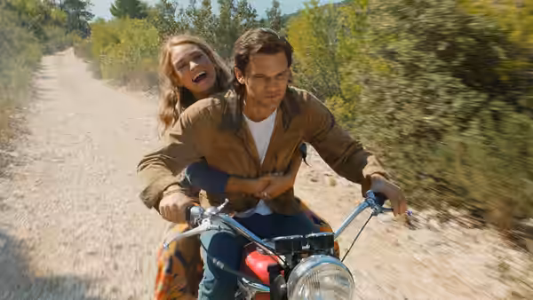 Young Donna (Lily James) rides behind a man on a motorcycle along a country road