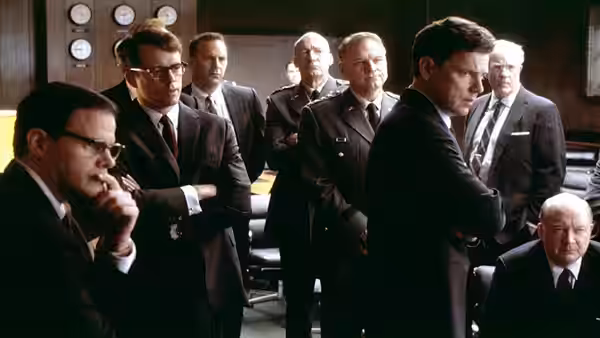 Kenneth O'Donnell (Kevin Costner) stands among officials gathered in a crowded meeting room