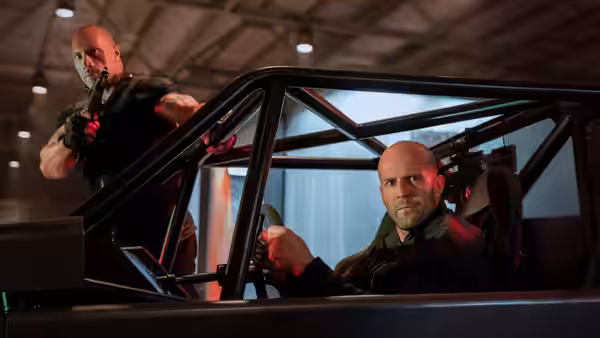Deckard Shaw (Jason Statham) drives a reinforced car as Luke Hobbs (Dwayne Johnson) rides beside him