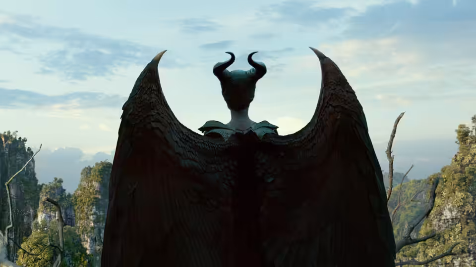 Maleficent (Angelina Jolie) faces a forested landscape, her enormous black wings spread wide
