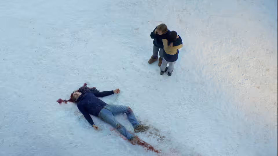 A body lies bleeding in the snow while a person stands nearby