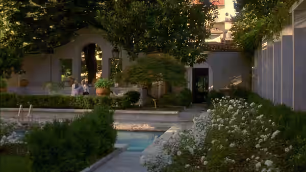Two people sit beside a swimming pool in a leafy courtyard garden
