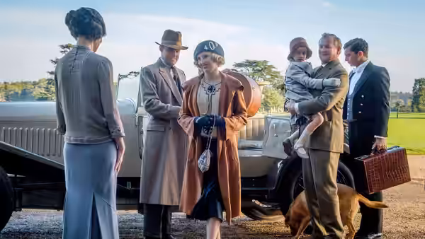 A woman in a cloche hat greets a group beside a parked car
