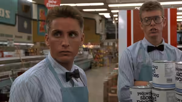 Otto Maddox (Emilio Estevez) stands wearing an apron in a supermarket