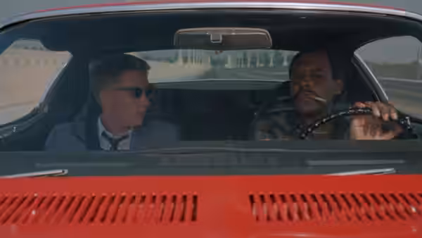 Two men talk while driving a red convertible along the highway