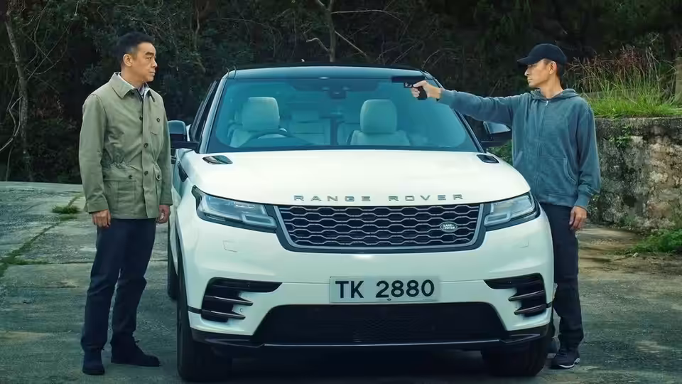 A man aims a pistol at another beside a white Range Rover on a wooded road