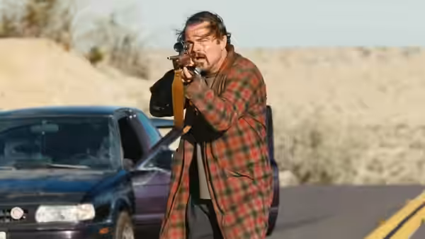 Bob (Leonardo DiCaprio) aims a rifle beside a car on a desert road