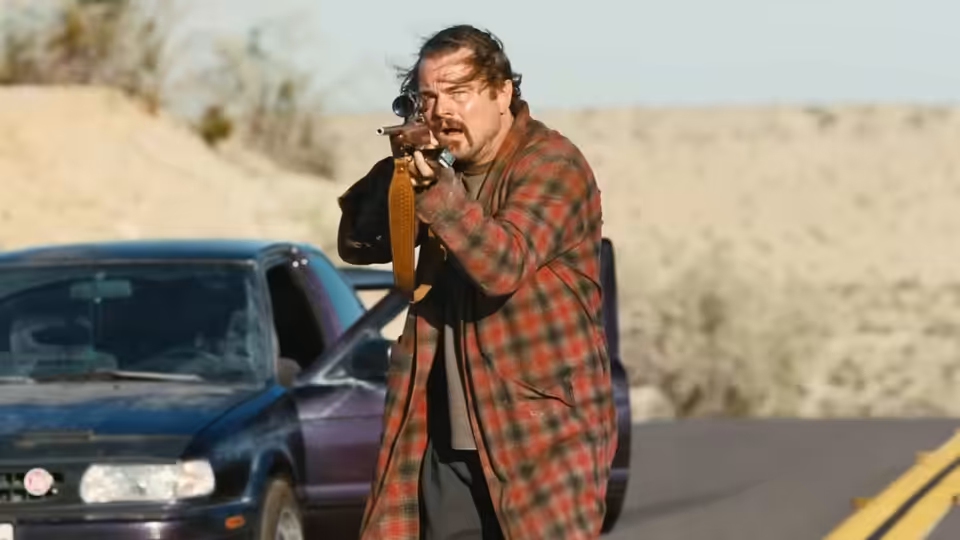 Bob (Leonardo DiCaprio) aims a rifle beside a car on a desert road
