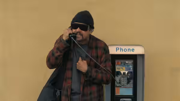 A man in a beanie talks on a payphone against a yellow wall