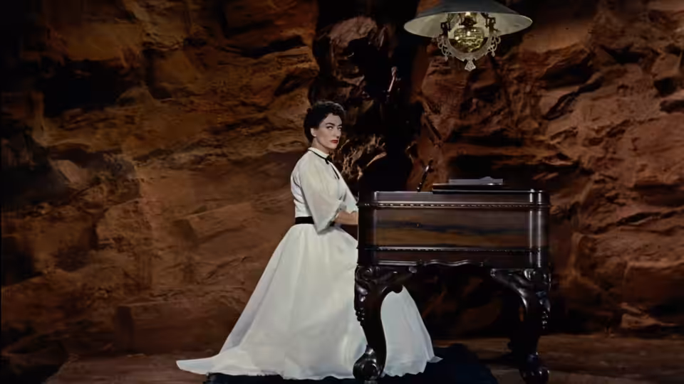 Vienna (Joan Crawford) stands beside a piano in a dimly lit room