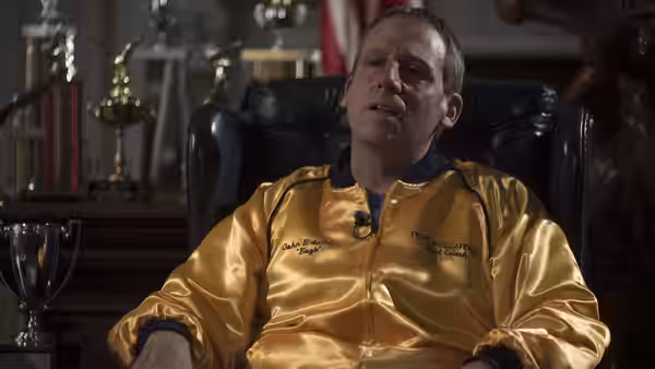 John du Pont (Steve Carell) sits in a leather chair wearing a gold jacket