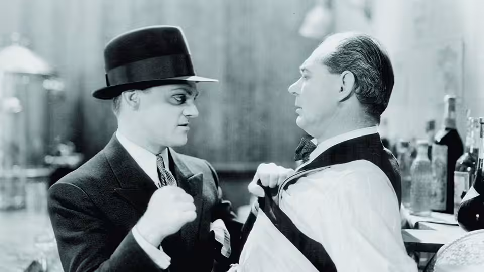 Two suited men face each other indoors, one gripping his lapel
