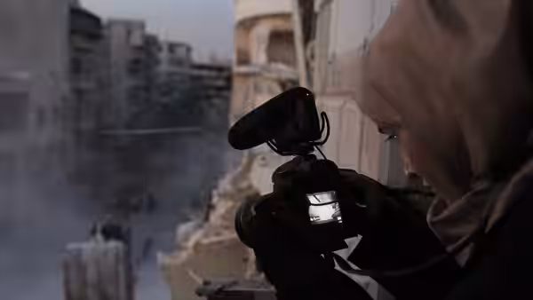 A woman in a headscarf films with a camera from a damaged building overlooking ruins