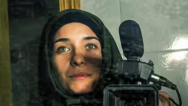 A woman in a green headscarf holds a video camera and looks forward