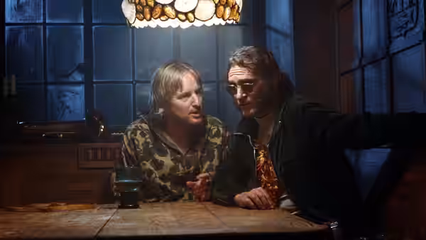 Doc Sportello (Joaquin Phoenix) leans across a table beside a man in a camouflage shirt