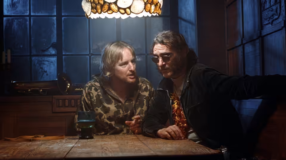 Doc Sportello (Joaquin Phoenix) leans across a table beside a man in a camouflage shirt