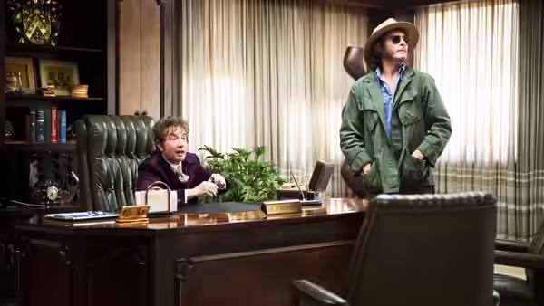 Doc Sportello (Joaquin Phoenix) stands in a private office beside a seated man behind a desk
