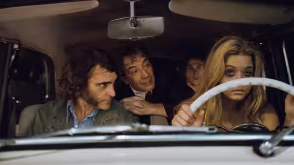 Doc Sportello (Joaquin Phoenix) sits beside a blonde woman driving, with two men in the back