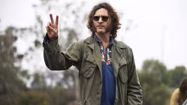 Doc Sportello (Joaquin Phoenix) raises two fingers outdoors, wearing sunglasses and a green jacket