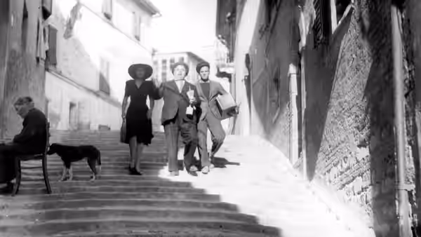 Three people descend a narrow street past a seated man and a dog