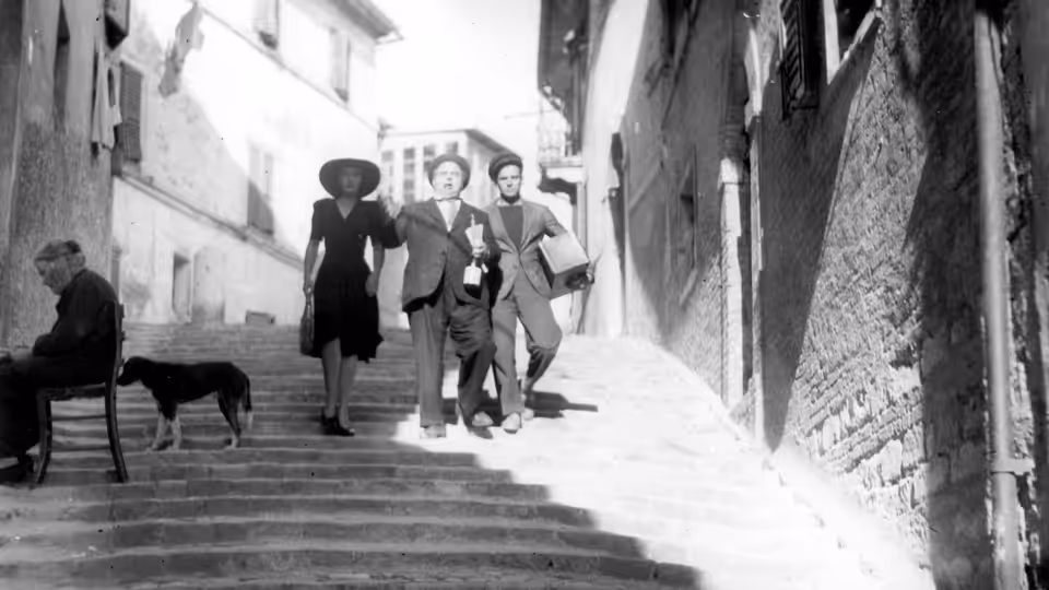 Three people descend a narrow street past a seated man and a dog