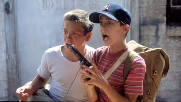 Two boys sit outside a building one holds a handgun in his mouth
