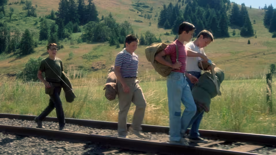 Four boys carrying duffel bags walk along railroad tracks in open countryside