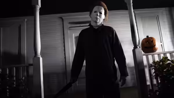 a masked figure in black stands on a porch holding a knife near a pumpkin