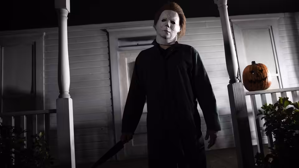 a masked figure in black stands on a porch holding a knife near a pumpkin