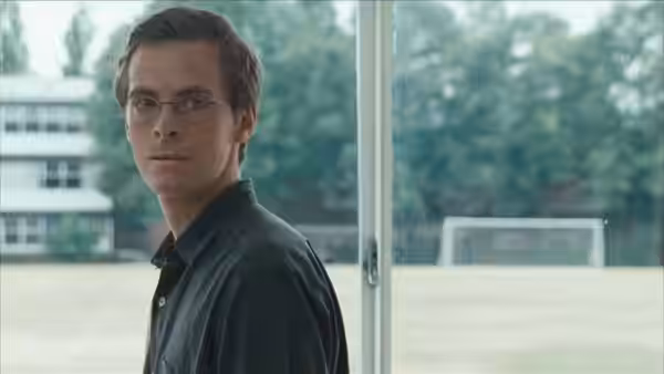 A bespectacled student stands by a window overlooking the school playing field