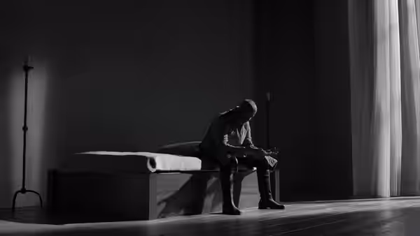 Macbeth seated on a bed in a dark room holding a blade