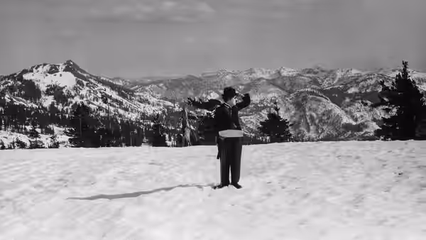 A lone man stands in a snowy field, facing a mountain range with his arms raised