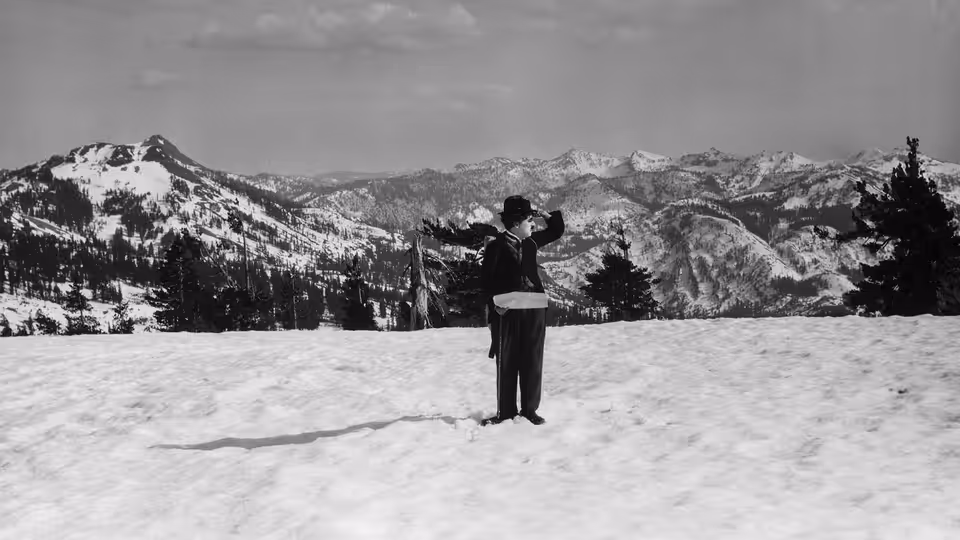 A lone man stands in a snowy field, facing a mountain range with his arms raised
