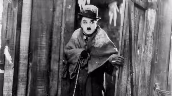 The Lone Prospector (Charlie Chaplin) peers through a doorway, wrapped in a blanket and holding his cane