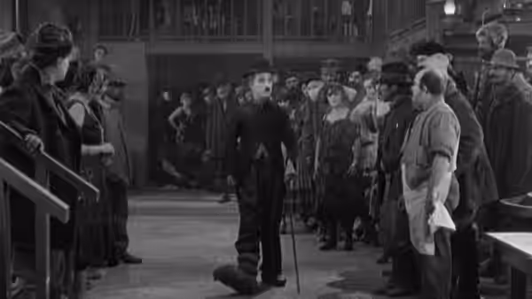 The Lone Prospector (Charlie Chaplin) stands with his cane among onlookers in a crowded room