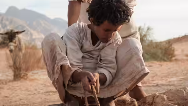 Theeb crouches to tie a rope around a stone as another person stands behind him