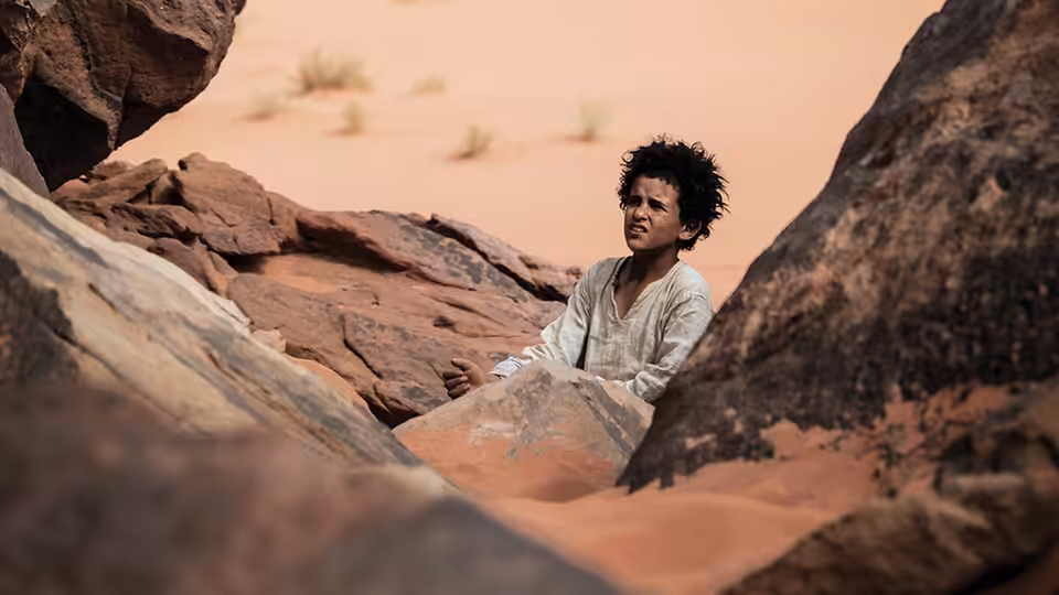 Theeb (Jacir Eid Al-Hwietat) sits among rocks in the desert