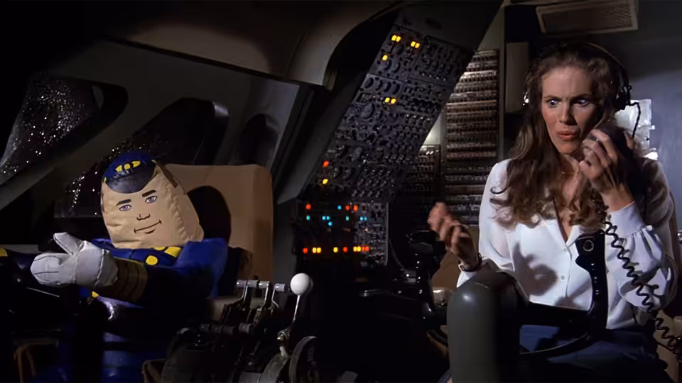 Woman in white shirt speaks on phone beside pilot dummy in cockpit