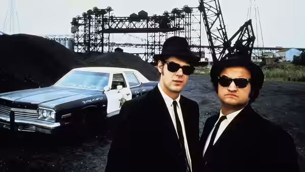 Elwood Blues (Dan Aykroyd) and Jake Blues (John Belushi) stand before a police car at an industrial yard