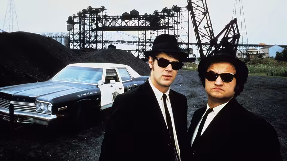 Elwood Blues (Dan Aykroyd) and Jake Blues (John Belushi) stand before a police car at an industrial yard
