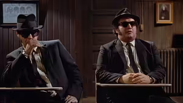 Jake Blues (John Belushi) and Elwood Blues (Dan Aykroyd) sit in a police interview room