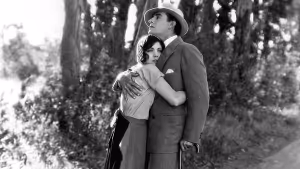A man in a suit and hat embraces a woman beneath the trees