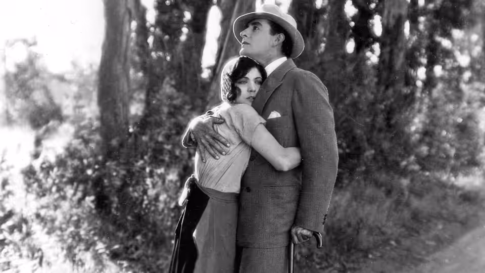 A man in a suit and hat embraces a woman beneath the trees